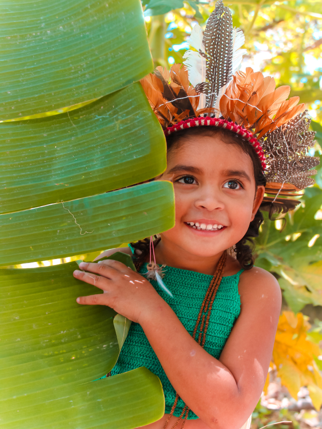 Criança indígena atrás de uma folha de bananeira usando um cocar feito de penas de aves laranja e cinza