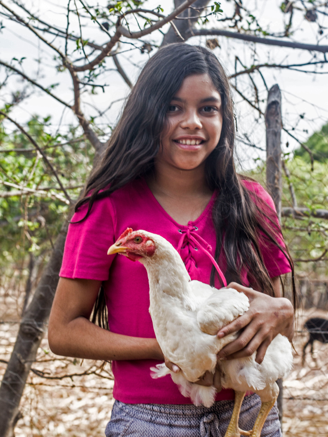 Menina de longos cabelos negros e lisos, pele parda, usando blusa cor de rosa. Ela sorri enquanto segura no colo uma galinha de penas brancas