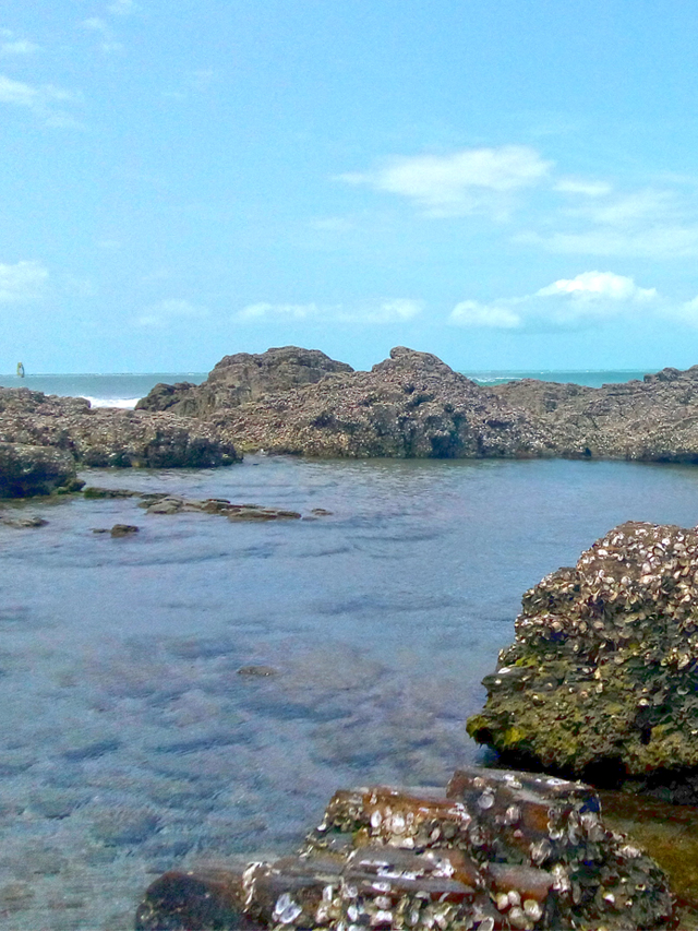Água do mar acumulada entre pedras na praia. Ao fundo, mar e céu azul
