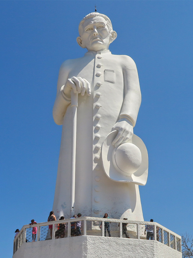 Estátua grande e branca da figura de um padre de frente, usando batina e segurando uma bengala e um chapéu. Aos pés da estátua há algumas pessoas visitando o monumento. Ao fundo céu azul