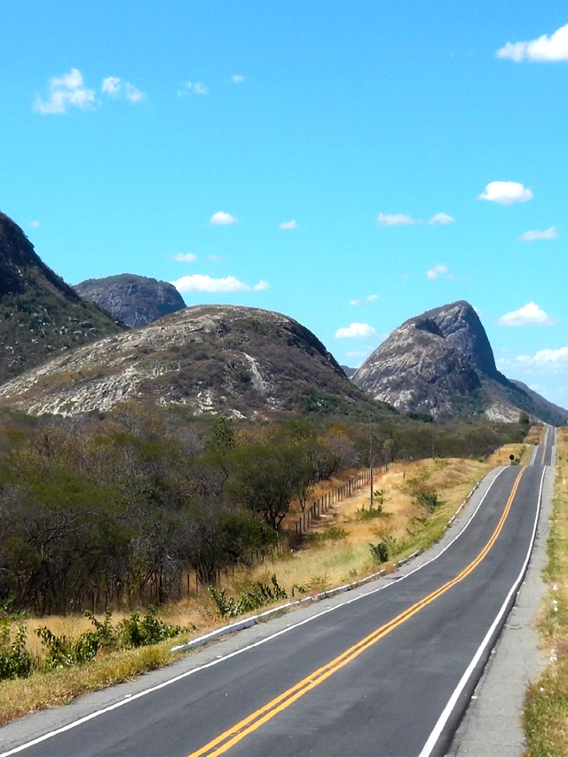 Estrada com grandes formações rochosas, como montanhas, do lado esquerdo, em meio à vegetação seca. Acima o céu azul com poucas nuvens