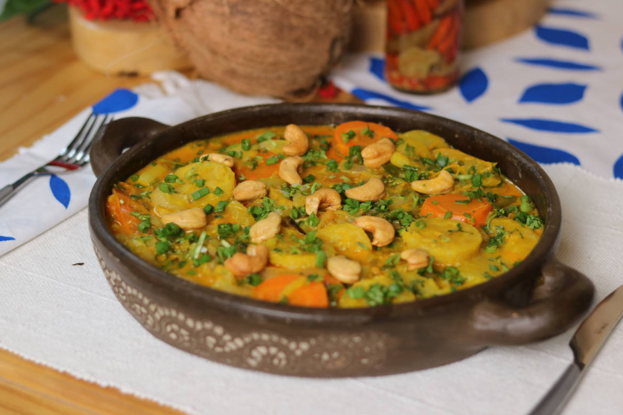 Panela rasa de barro com duas alças com um cozido de coloração amarela e castanhas por cima sobre um jogo americano cru com um garfo sobre um guardanapo branco com folhas azuis do lado esquerdo e uma faca do lado direito