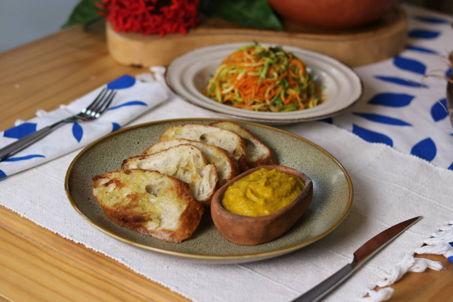 Prato rústico bege com cinco torradas do lado esquerdo e uma pequena travessa de barro com um creme amarelo do lado dieito sobre um jogo americano cru com um garfo sobre um guardanapo branco com folhas azuis do lado esquerdo e uma faca do lado direito. À frente, do lado esquerdo, fora de foco, outro prato com macarrão de legumes