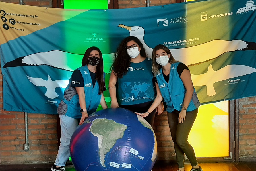 Três jovens do sexo feminino, usando máscaras, duas com colete e uma com camiseta do Projeto Albatroz azuis por trás de um globo terrestre inflável tendo ao fundo uma grande bandeira do Projeto Albatroz à frente de uma parede de tijolinhos vermelhos