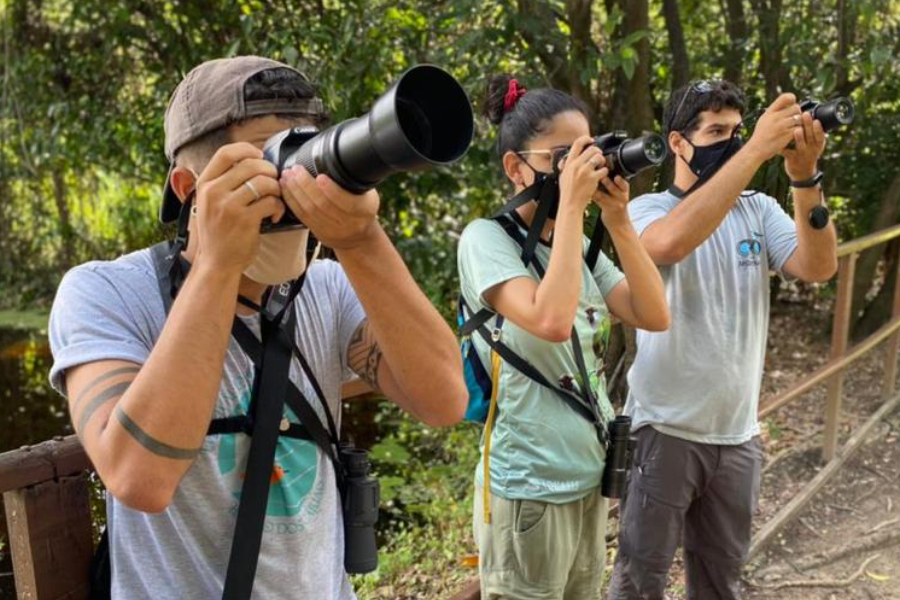 Três pessoas, dois homens nas pontas e uma mulher ao centro, usando máscaras e fotografando