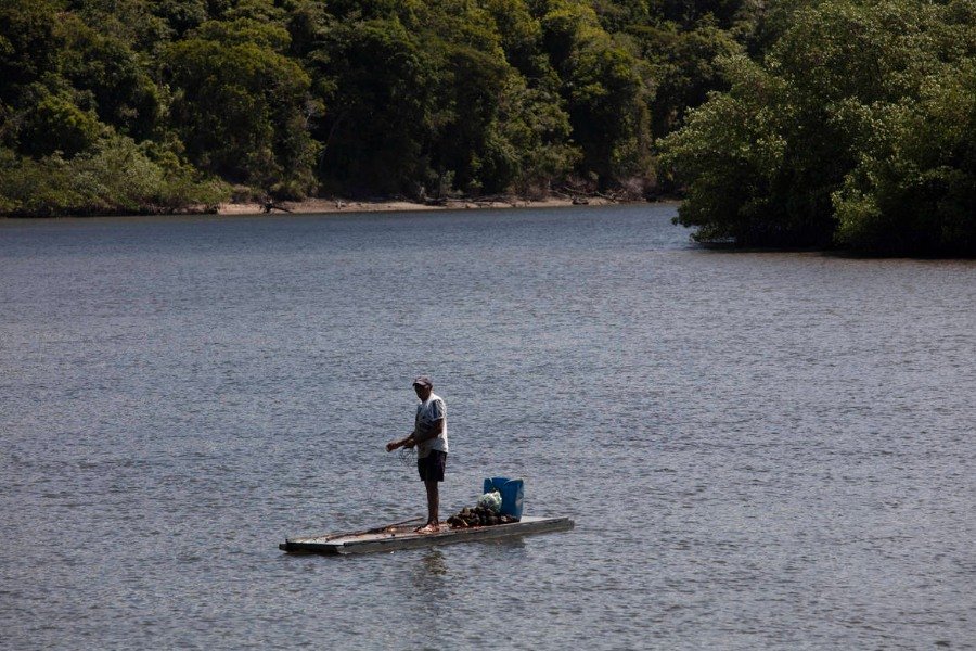 Pescador em pé em pequena embarcação no meio de um rio com vegetação de mangue às margens