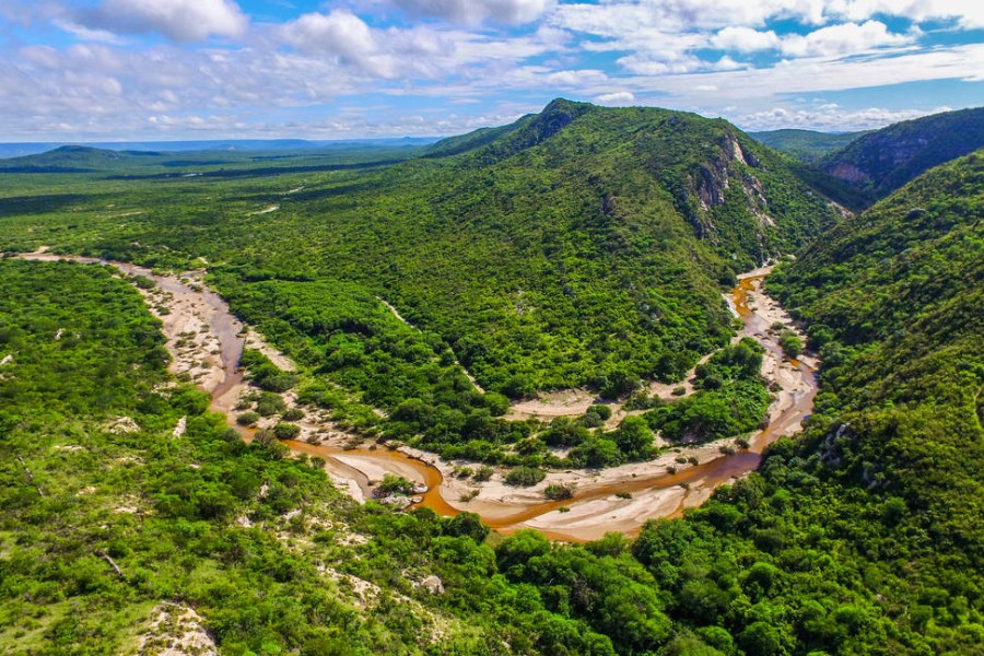 Vale e montanhas verdes com um rio meio seco que serpenteia entre elas. Ao fundo céu azul com nuvens brancas