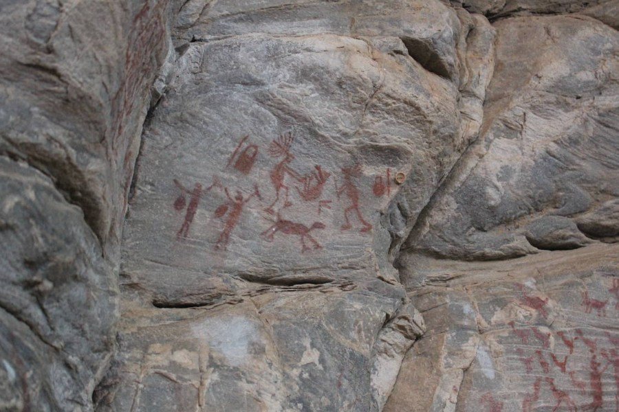 Rocha com inscrições rupestres de tonalidade marrom representando quatro pessoas caçando e abatendo animais e seus utensílios