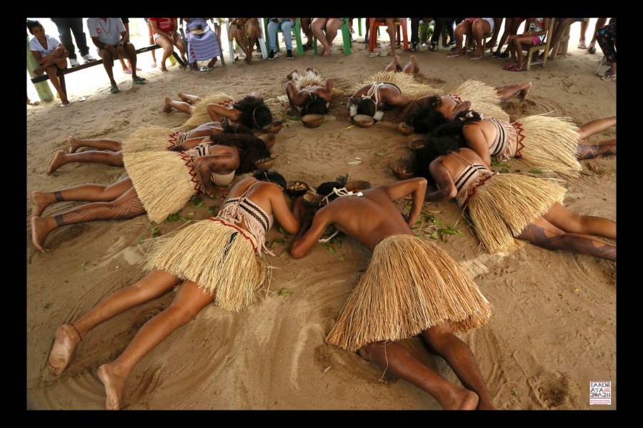 Dez indígenas trajados com saias de palha deitados de bruços na areia em forma circular com cabaças contendo alimentos à sua frente. Ao fundo é possível ver as pernas de pessoas sentadas em bancos e cadeiras acompanhando o ritual
