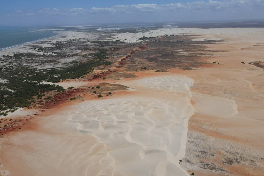 Imagem aérea de terreno avermelhado com dunas de areia clarinha ao centro e vegetação em direção à praia, onde a areia clara ressurge