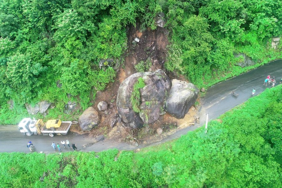 Vista aérea frontal de área verde cortada por pista com enormes blocos rochosos deslizados sobre ela. À esquerda há sete pessoas em pé próximas a uma carreta com equipamento sobre ela