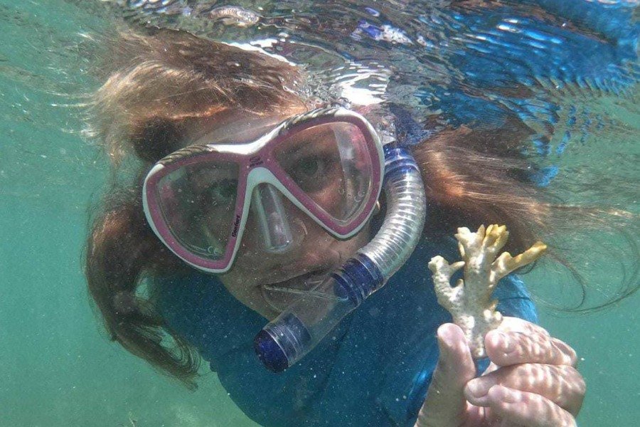 Embaixo d'água, mulher branca de longos cabelos ruivos usando máscara de mergulho branca e rosa com respirador azul e transparente e camiseta azul segura fragmento de coral branqueado