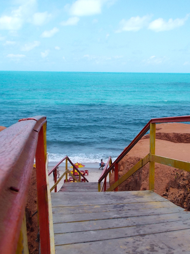 Escadaria que liga o alto da falésia alaranjada à areia da praia. Os corrimões da escada são de madeira pintados de laranja e amarelo. Ao fundo mar e céu azuis