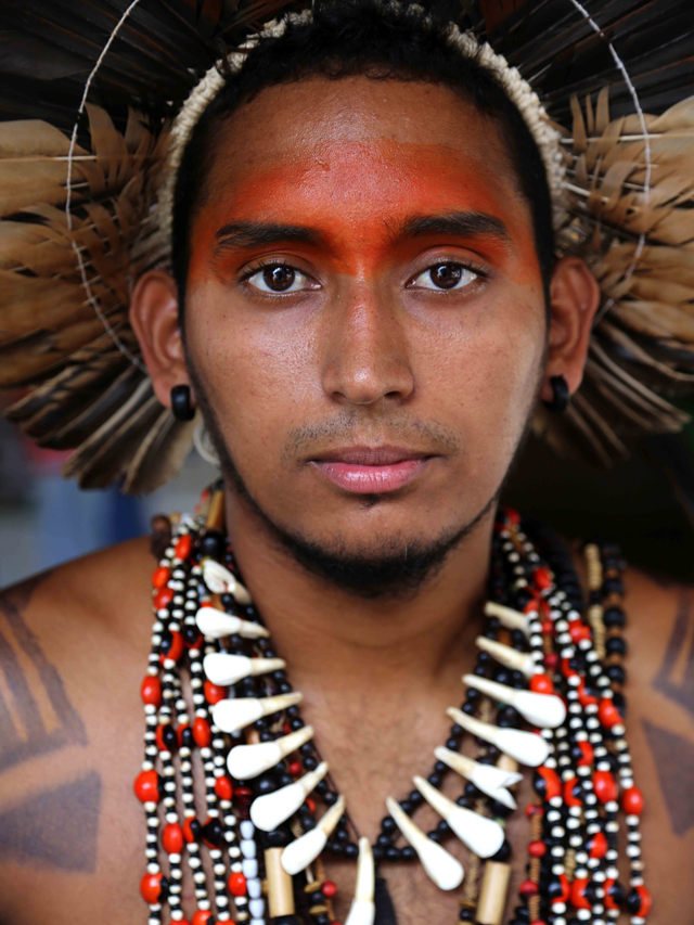 Homem indígena, usando cocar feito de penas. Ele tem a testa pintada de vermelho e pinturas corporais pretas na altura dos ombros. O indígena usa colares feitos de sementes e dentes de animais