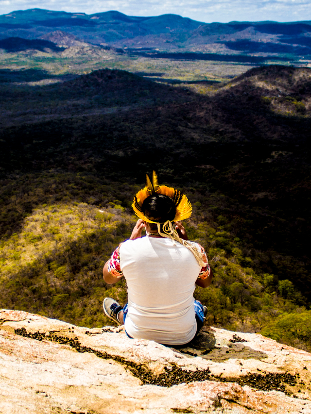 Indígena homem sentado em um mirante com vista para grande área de vegetação e montanhas. Ele usa um cocar de penas de aves amarelas e está sentado de costas