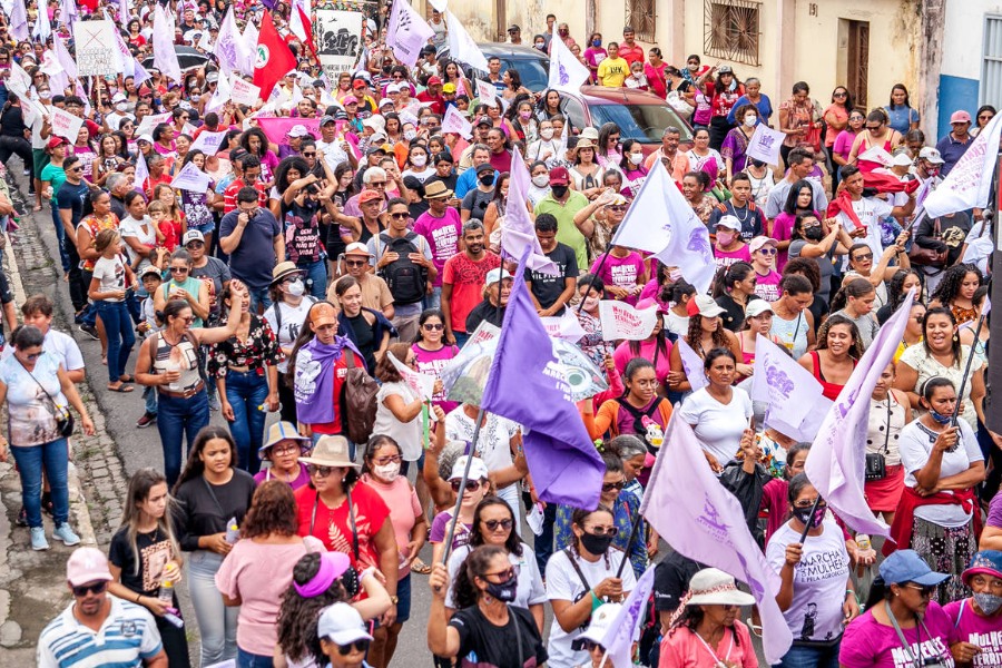 centenas de mulheres vestindo branco, vermelho e lilás predominantemente, com algumas bandeiras nessas cores, marcham por uma rua