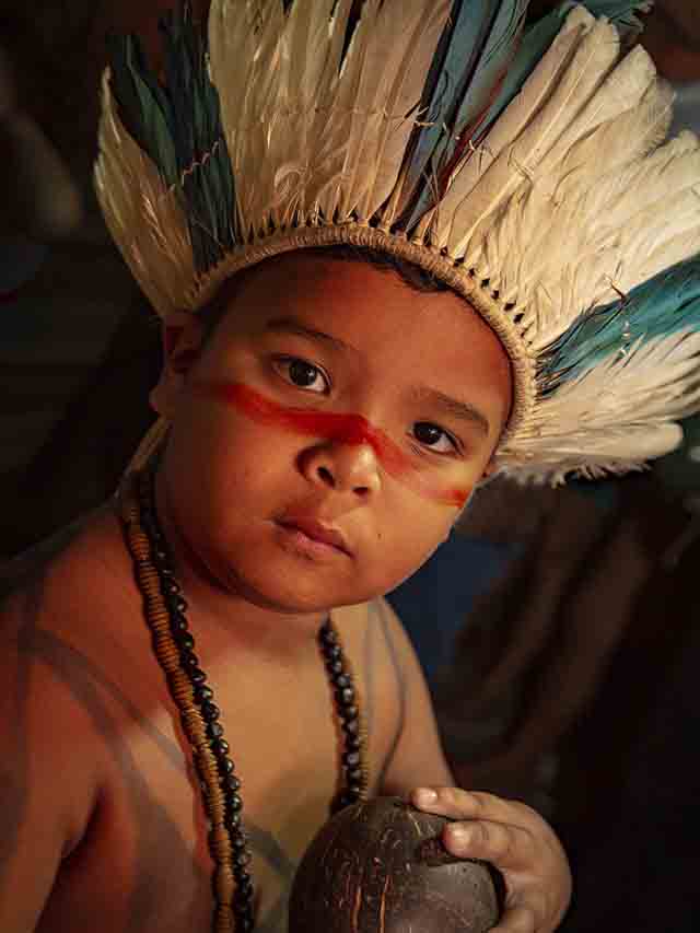 Retrato de criança Indígena (Saulo), com o rosto pintado de vermelho, usando um cocar de penas de aves brancas e azuis e colares feitos de sementes