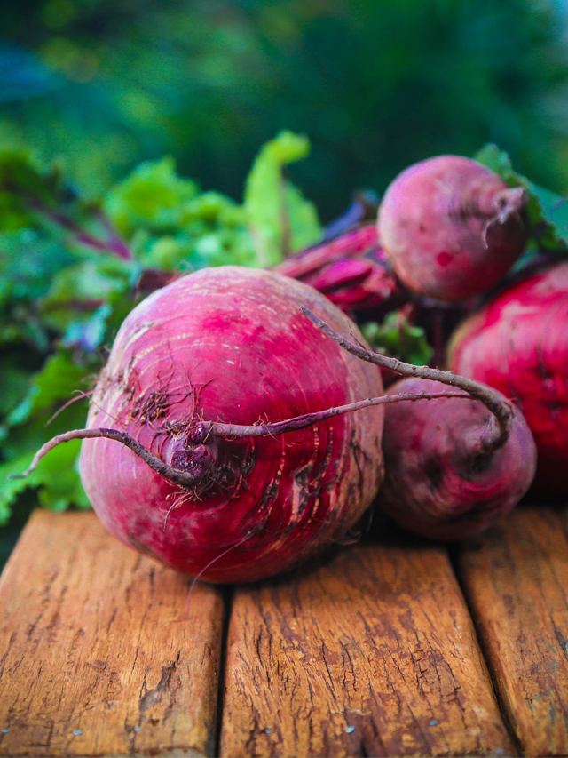 quatro beterrabas de tamanhos diferentes e de cores avermelhadas com talos verdes, dispostas em cima de uma superfície de madeira
