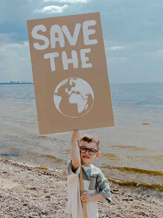 Fotografia de menino branco usa óculos, bermuda cinza e camisa branca com detalhes cinza. Ele está em uma praia e segura uma placa de papelão na cor natural, bege, pintada de branco com os dizeres ‘SAVE THE’, em português ‘SALVE O’ e o desenho do Planeta Terra