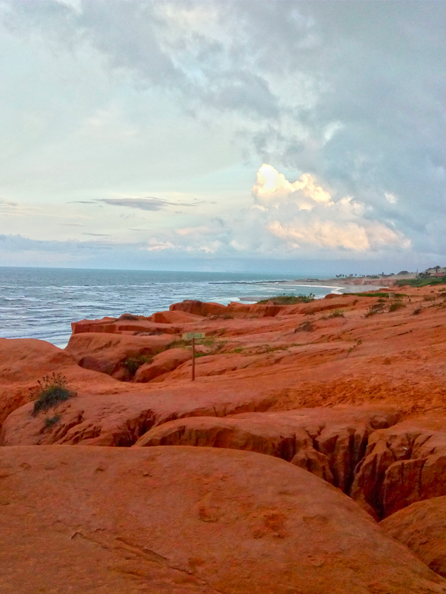 Fotografia de praia com céu nublado e raios de sol entre as nuvens. Mar ao fundo e altas falésias de coloração alaranjada vistas do topo