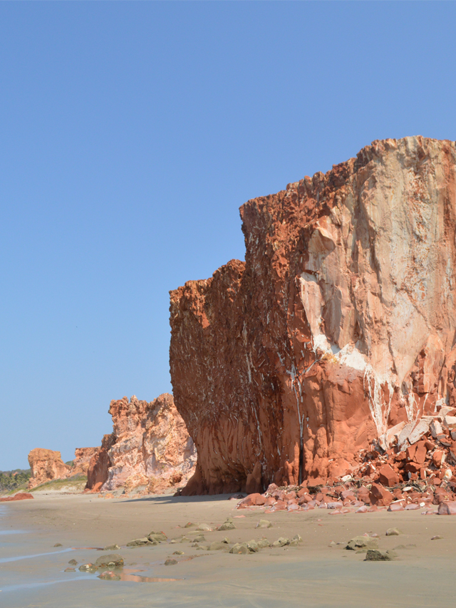 Fotografia de praia com uma grande falésia de coloração avermelhada à direita, a areia à esquerda, um pedacinho do mar à esquerda céu azul