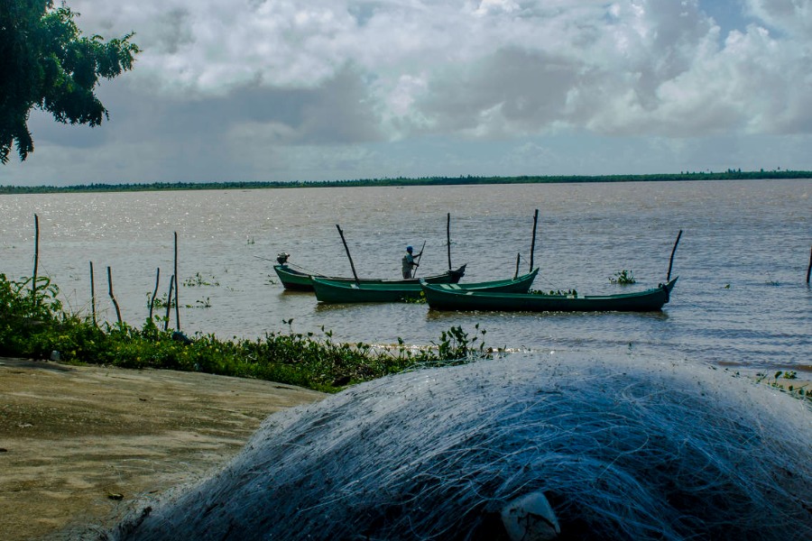 No primeiro plano há uma rede de pesca branca dobrada. Ao fundo, três barcos verdes à beira de um grande espelho d’água. Num deles há um homem em pé. No alto, céu nublado