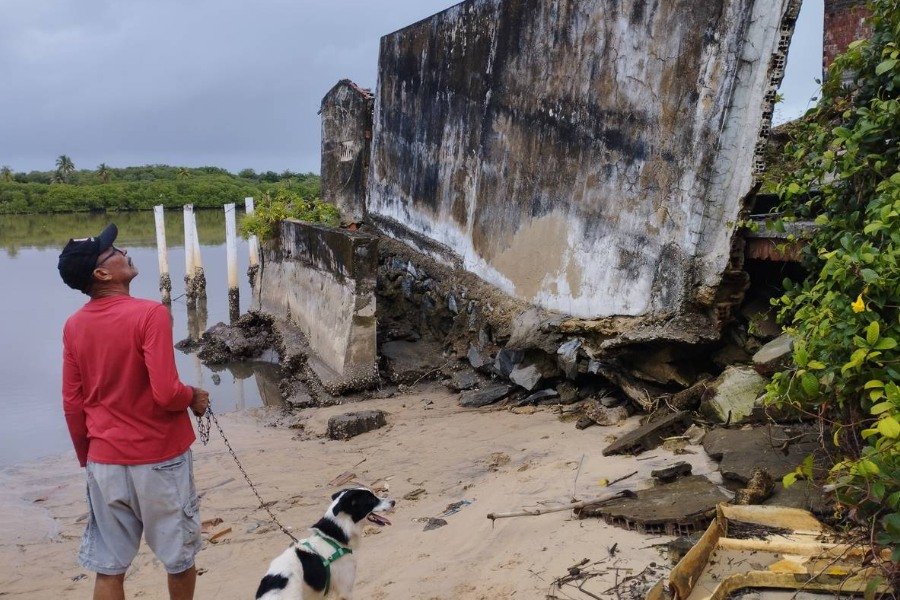 Homem pardo observa ruínas em beira de rio. Ele segura corrente atada a cachorro branco com pintas pretas