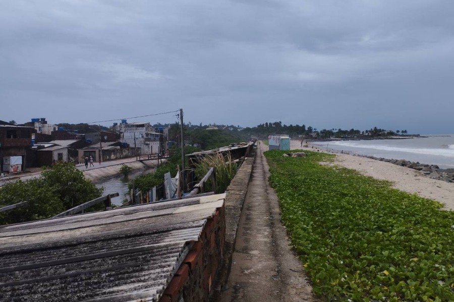 Foto de perspectiva de paredão. À esquerda, comunidade e canal. À direita, vegetação rasteira, areia e mar. Acima, céu nublado