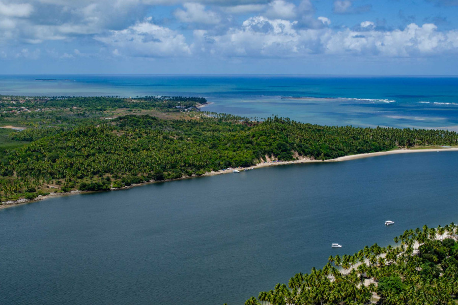 Fotografia de Paisagem de encontro de rio com o mar com águas de coloração predominantemente azulada com uma faixa de terra coberta de vegetação onde se destacam coqueiros e praias de areia branca às margens