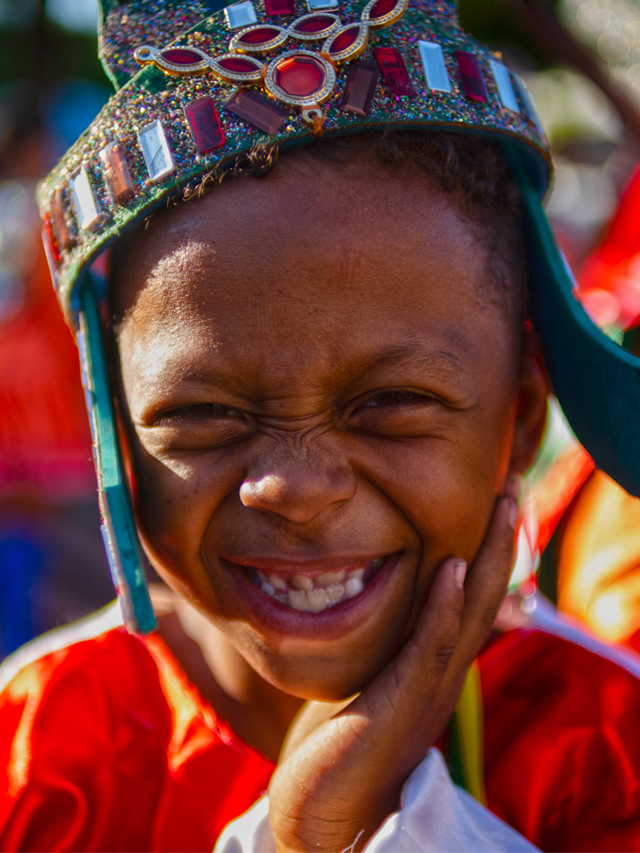 Fotografia de Menino negro com uma das mãos no queixo enquanto sorri. A criança usa roupas vermelhas e uma coroa de EVA azul com glitter e pedrarias falsas coloridas