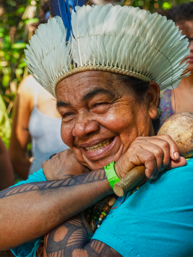 Fotografia de uma senhora indígena, usando um cocar de penas de aves brancas e azuis, de cabelos negros. A mulher usa camiseta azul celeste e colares feitos com sementes marrons e vermelhas. Ela sorri e tem os braços com pinturas étnicas. Os braços dela estão cruzados como se estivesse abraçando algo e em uma das mãos ela possui um chocalho marrom feito de cabaça