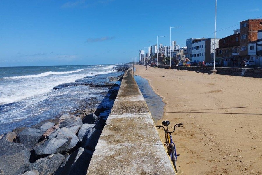 Foto de perspectiva de paredão com pedras e mar à esquerda, areia, rua e construções à direita. Ao fundo, céu azul. Há uma bicicleta encostada no paredão