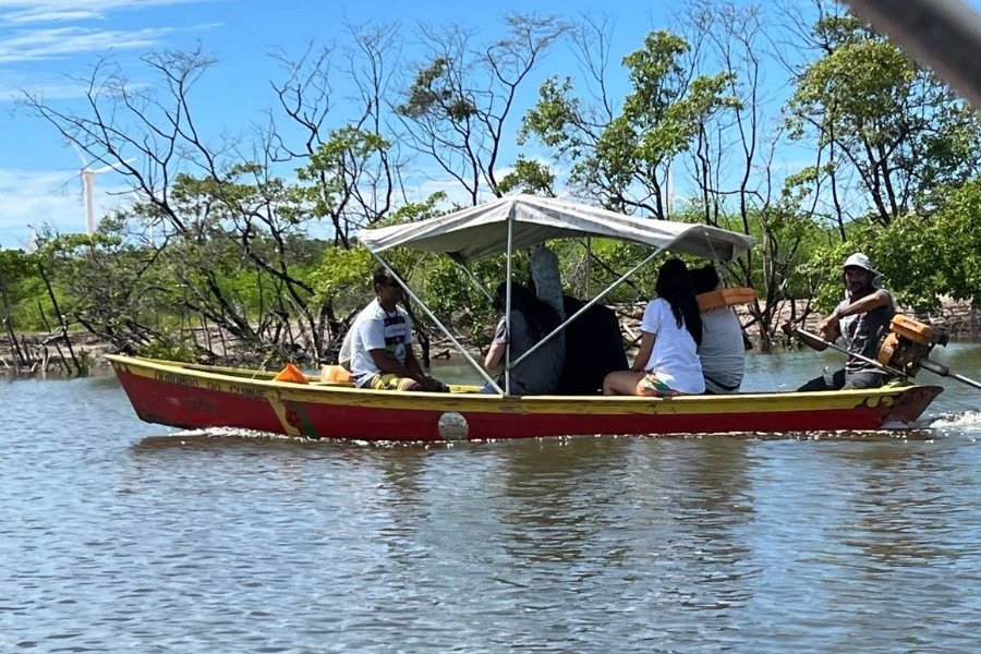 Barco vermelho e amarelo com uma coberta branca de lona e um pequeno grupo de pessoas nele navegando um rio de águas escuras. Ao fundo, vegetação rala, céu azul e um aerogerador do lado esquerdo