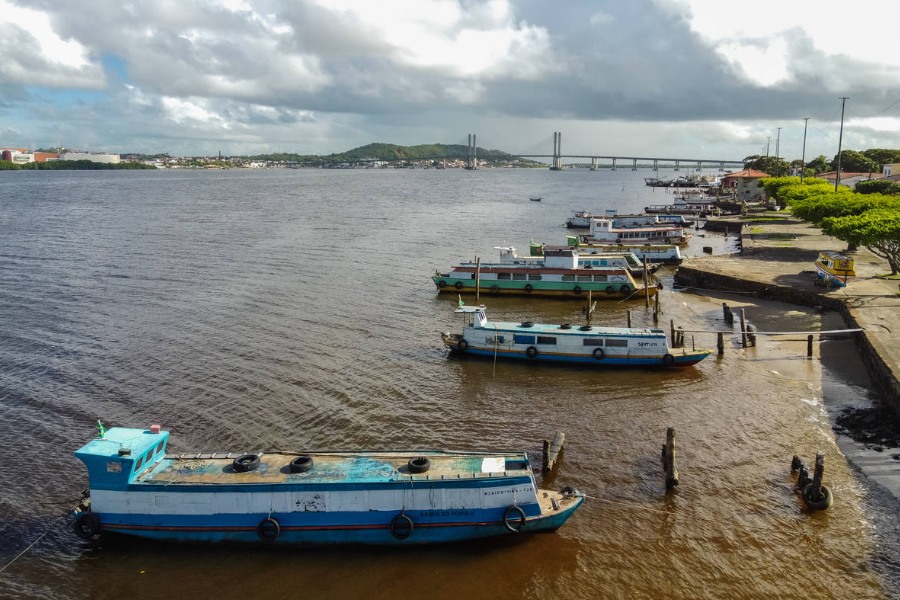 Fotografia de embarcações pequenas ancoradas nas margens de um largo rio com uma grande ponte que o cruza ao fundo sob céu azul com nuvens brancas