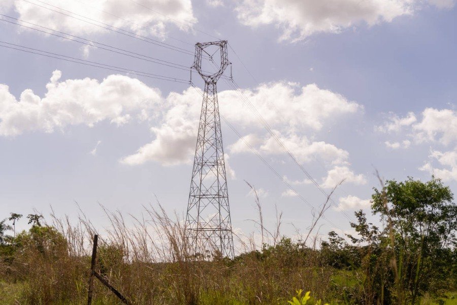 Fotografia de céu azul com nuvens, vegetação rasteira e, no meio da paisagem, uma grande estrutura de ferro com fios, uma torre que funciona como linha de transmissão de energia elétrica