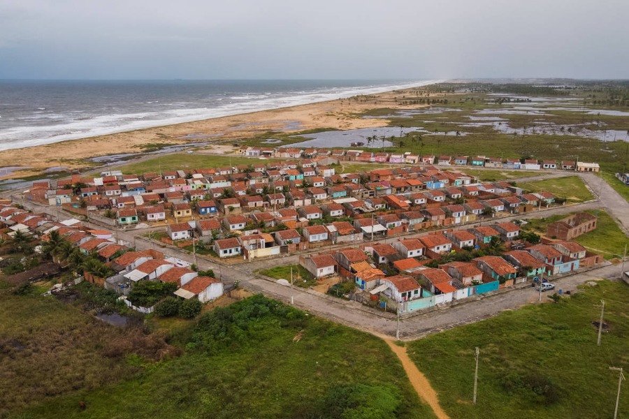 Fotografia aérea de ruas e casinhas enfileiradas em comunidade ao lado da praia, ao fundo. A comunidade está cercada por vegetação e lagoas sob contém céu azul com nuvens brancas