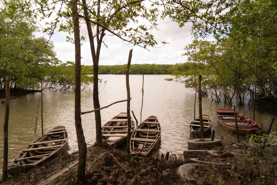 Fotografia de céu com nuvens brancas, vegetação de mangue ao fundo, grande espelho d'água de rio, canoas às margens de um espelho de água