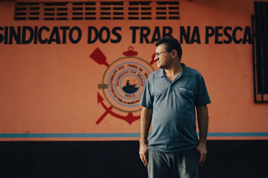Fotografia de homem em pé, de cabelos negros, usando óculos, vestindo camisa cinza e bermuda da nesma cor. Ele está em frente a uma parede laranja, onde está escrito SINDICATO DOS TRAB NA PESCA