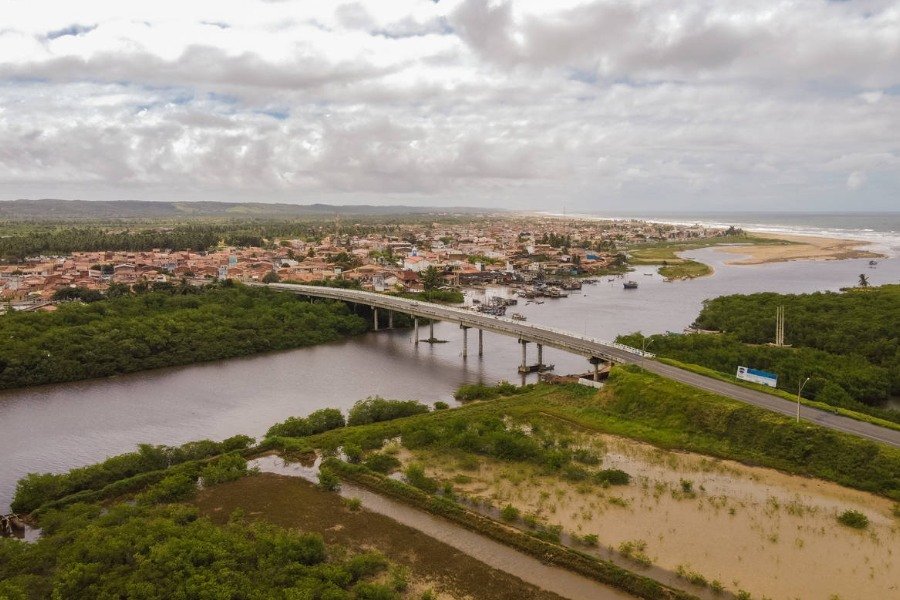 Fotografia aérea de grande ponte cruzando um rio que se encontra com o mar, no canto direito da imagem. Na margem direita há vegetação verde e áreas alagadas e na esquerda há construções e casas. Ao fundo, céu azul com nuvens brancas