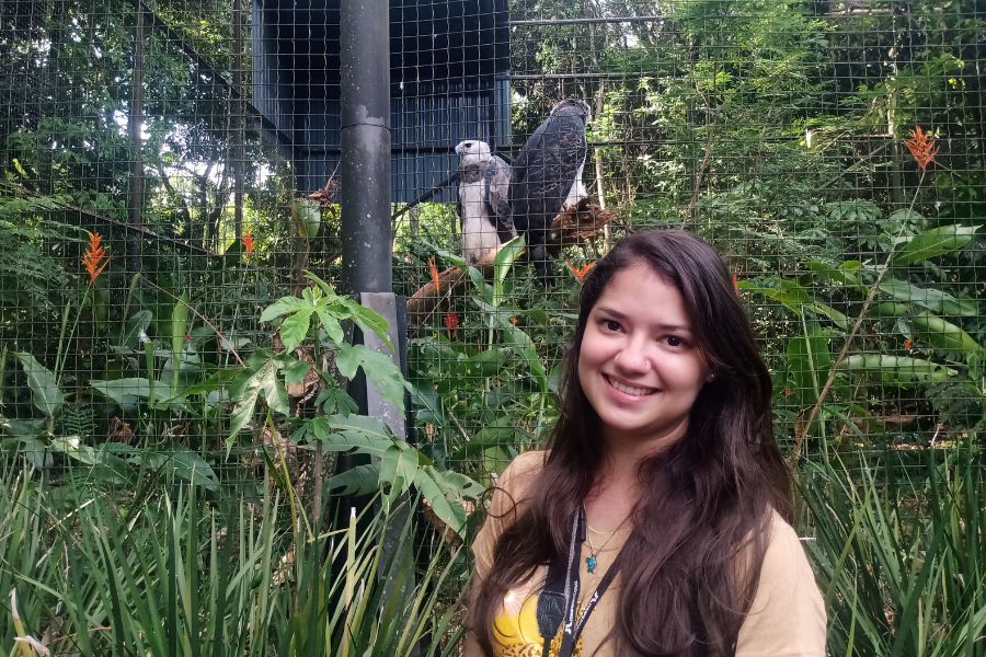 Mulher branca de cabelos escuros, lisos e longos sorri para a foto. Atrás dela um viveiro com grandes aves brancas e pretas