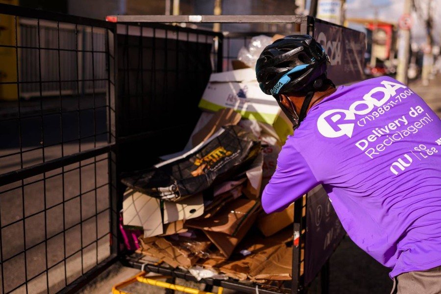Homem negro usando camiseta lilás com a logo marca RODA, número de telefone e Delivery da Reciclagem! e capacete de ciclista preto arruma materiais recicláveis numa estrutura metálica na traseira de uma bicicleta