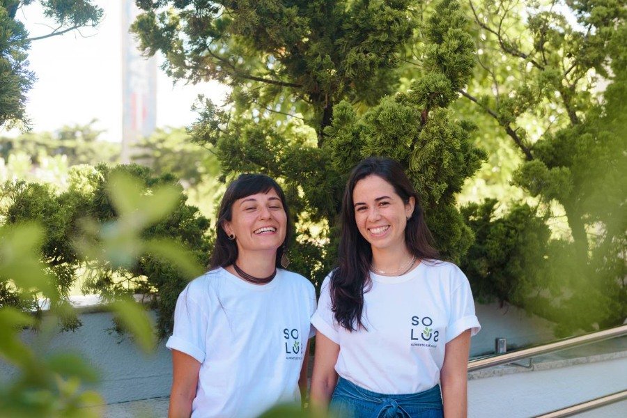 Duas mulheres brancas e sorridentes posam para foto com camisetas brancas com a logomarca SOLOS tendo ao fundo copas de árvores verdes