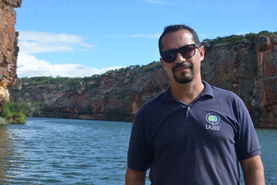 Fotografia de homem de cabelos negros e cavanhaque, usando óculos escuros e uma camisa azul marinho. Ao fundo dele há uma paisagem de grandes paredões de rochas alaranjadas por onde passa um caudaloso rio