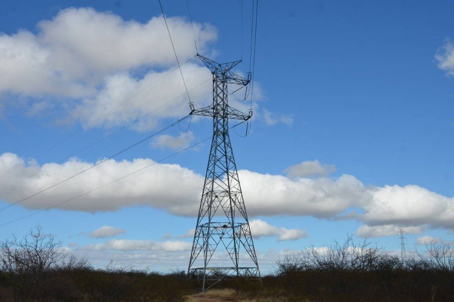 Torre de linha de transmissão de energia elétrica em uma área de vegetação seca e rasteira sob céu azul com nuvens