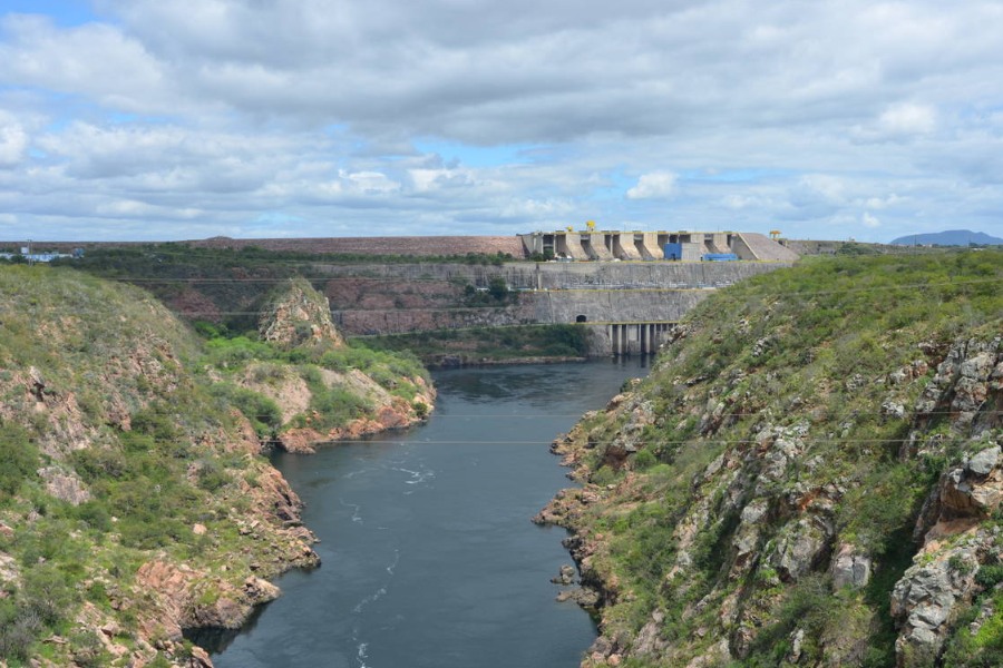 Grandes paredões de rochas cobertos por vegetação ladeiam um espelho d'água. Ao fundo há uma barragem de concreto sob um céu azul