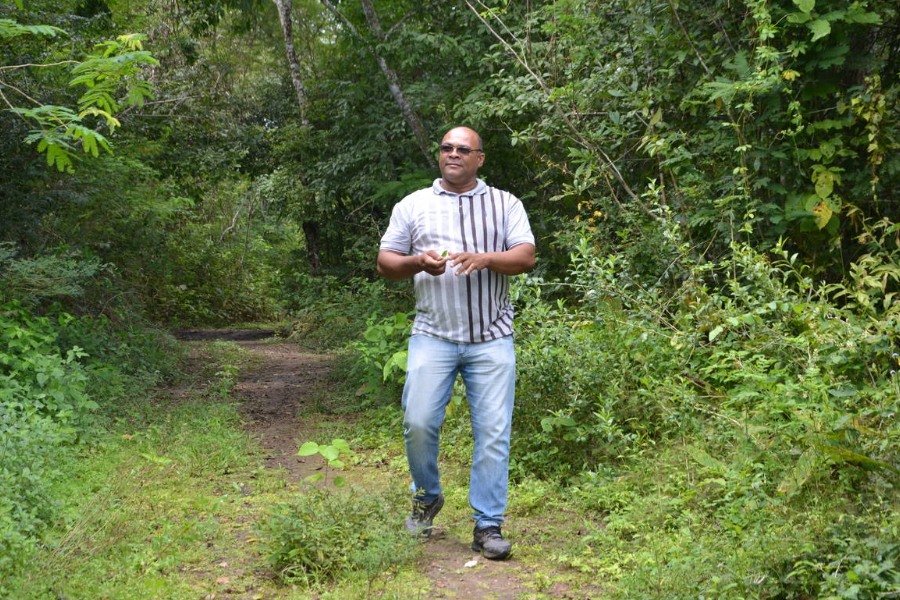 Fotografia de homem alto, careca, de pele negra. Ele usa óculos, camisa cinza e calça jeans e caminha numa trilha em meio à vegetação