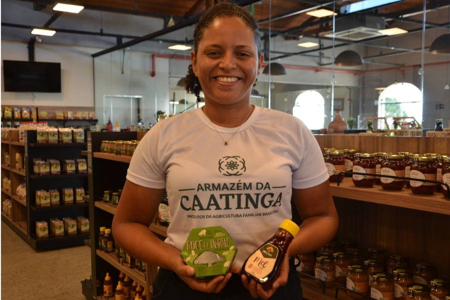 Mulher negra de cabelos cacheados negros presos, sorrindo enquanto segura uma caixa verde e um frasco marrom. Ela está entre prateleiras de produtos alimentícios e usa uma camisa branca com os dizeres "Armazém da Caatinga"