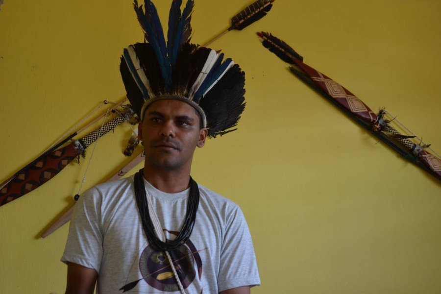 Fotografia de homem indígena, usando camisa branca e colar feito de sementes. Ele usa também um cocar feito em penas de aves azuis e brancas. Atrás dele há uma parede amarela onde estão pendurados artefatos povo semelhantes a flechas