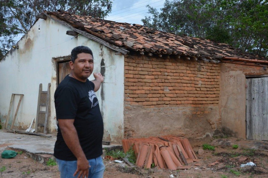 Homem indígena vestido de camisa preta e calça jeans apontando para uma casa de fachada branca, telhas e tijolos marrons e telhas encostadas em uma de suas paredes