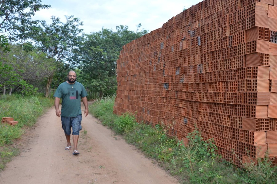 Homem indígena vestido com camisa e bermuda cinzas caminhando em uma estrada ladeada por vegetação verde. Ao lado direito há uma pilha de tijolos marrons